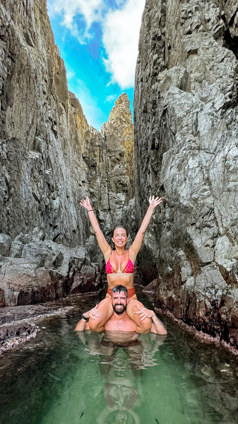 Agnes de braços erguidos sobre Thiago em uma piscina natural entre as pedras