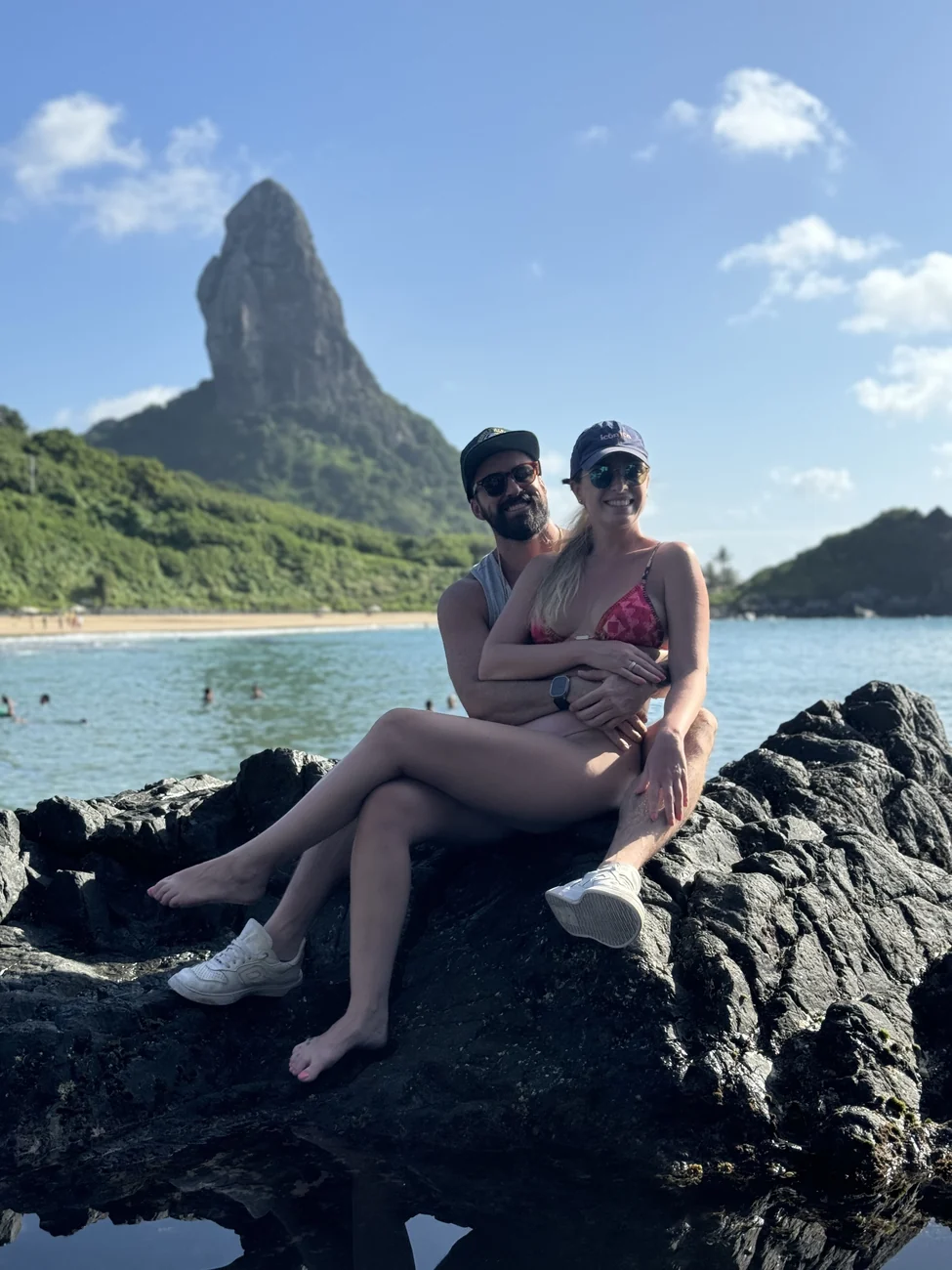 O casal sentado em uma pedra com o morro e o mar ao fundo