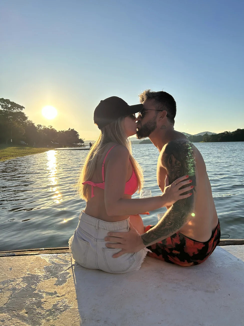 Agnes e Thiago se beijando ao pôr do sol, à beira da água